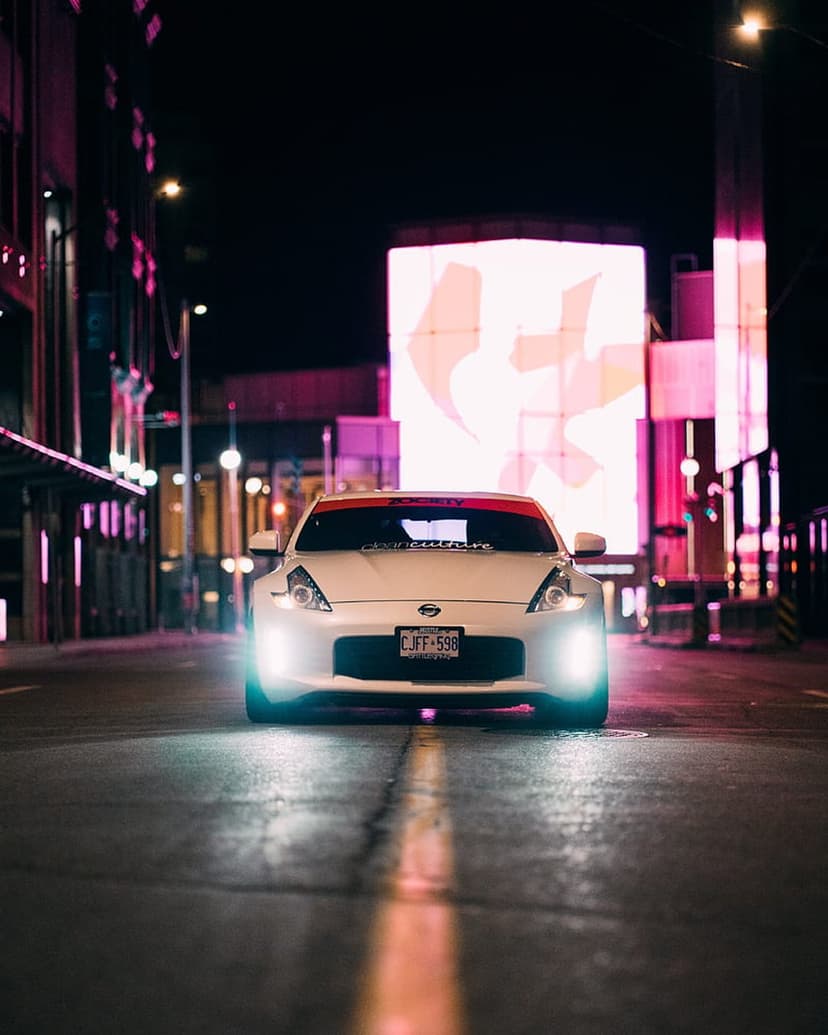White Nissan 370Z with lit headlights on a damp city street framed by vibrant neon skyscrapers at night.
