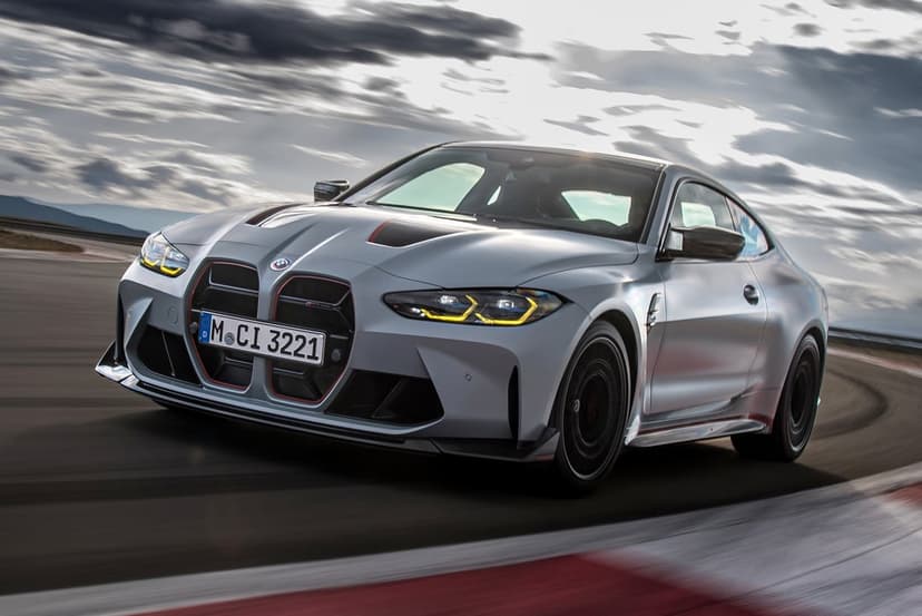 Silver BMW M4 CSL with yellow headlights and red accents driving fast on a racetrack under a cloudy sky.