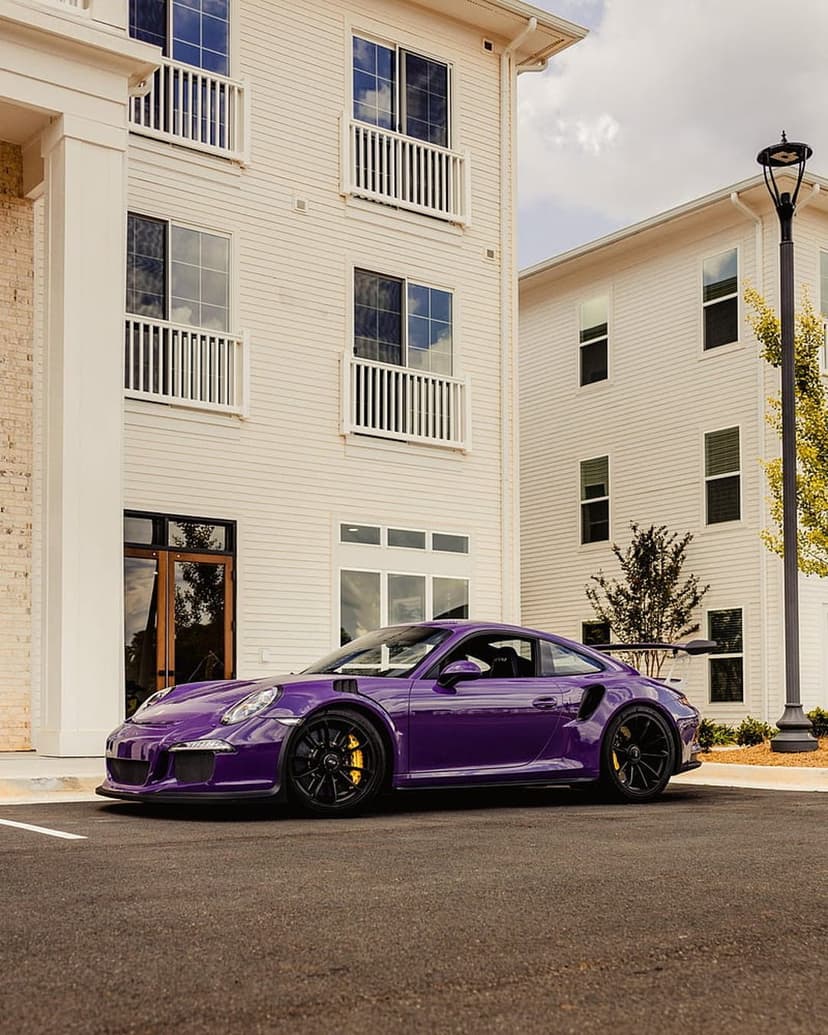 Purple Porsche GT3 RS with black wheels parked in front of an off-white modern building with glass balconies.