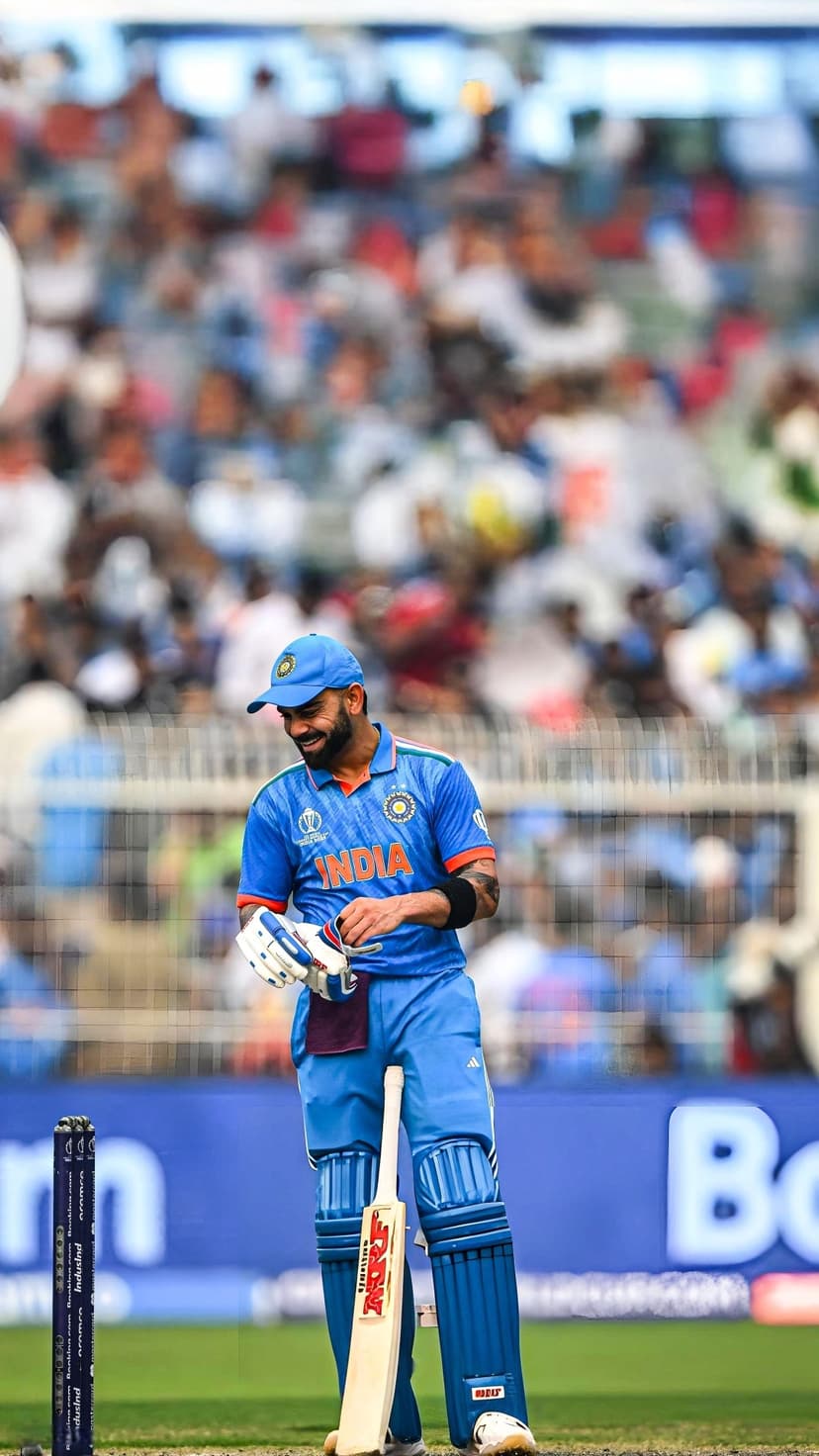 Virat Kohli smiles in a blue India kit beside a cricket bat on a field, framed for a vertical mobile wallpaper.