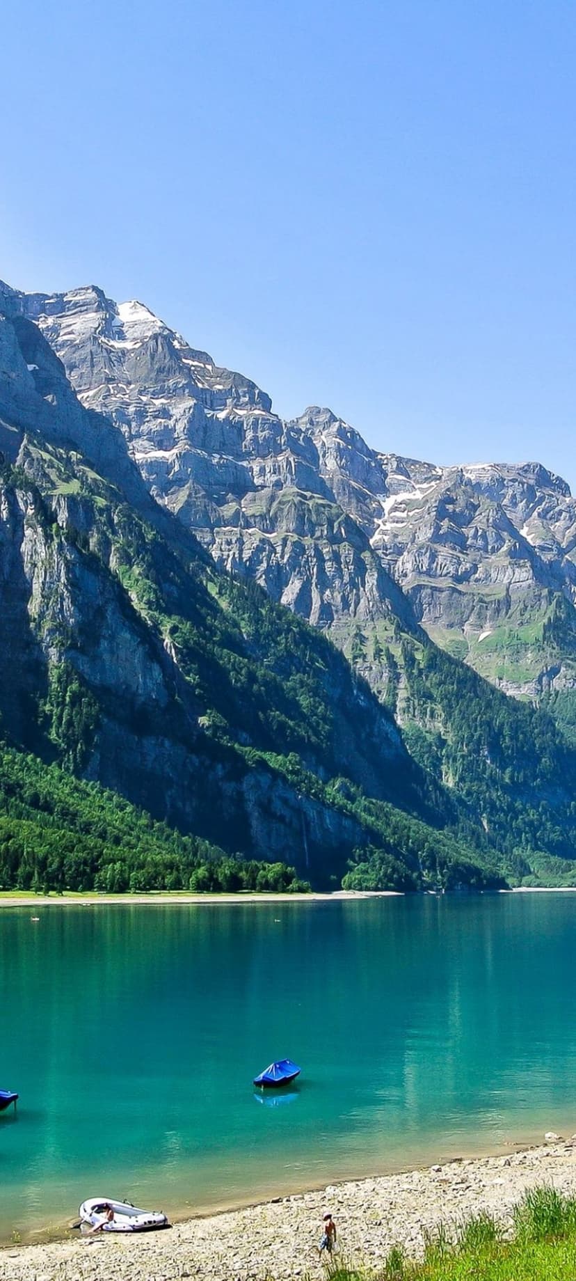 Vibrant teal lake with wooden boats backed by snow-dusted peaks and pine forests under a clear blue summer sky.