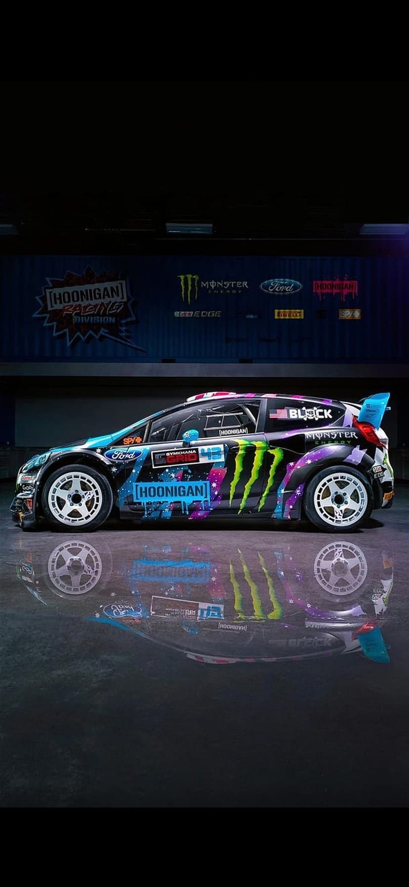 Hoonigan Ford Fiesta rally car with Monster Energy livery reflecting on a glossy dark floor at a sharp angle.