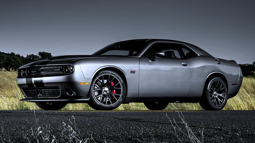 Silver Dodge Challenger muscle car with black racing stripes on an asphalt road against a moody sky.