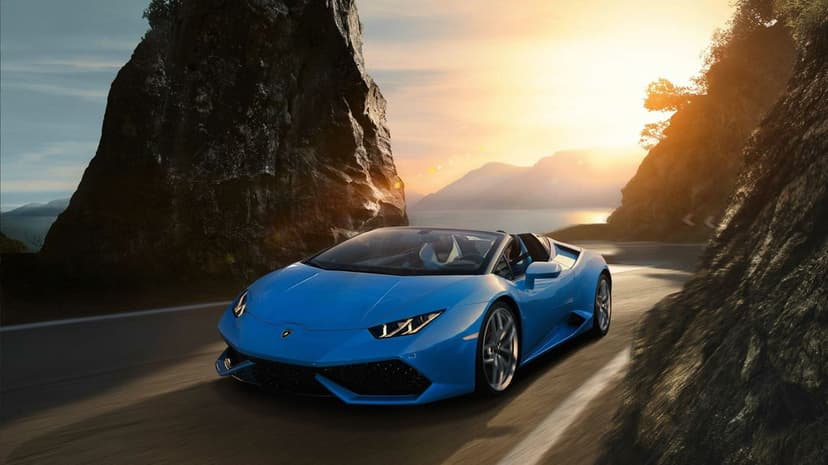 Blue Lamborghini convertible driving on a winding road between high cliffs at sunset with mountain vistas.
