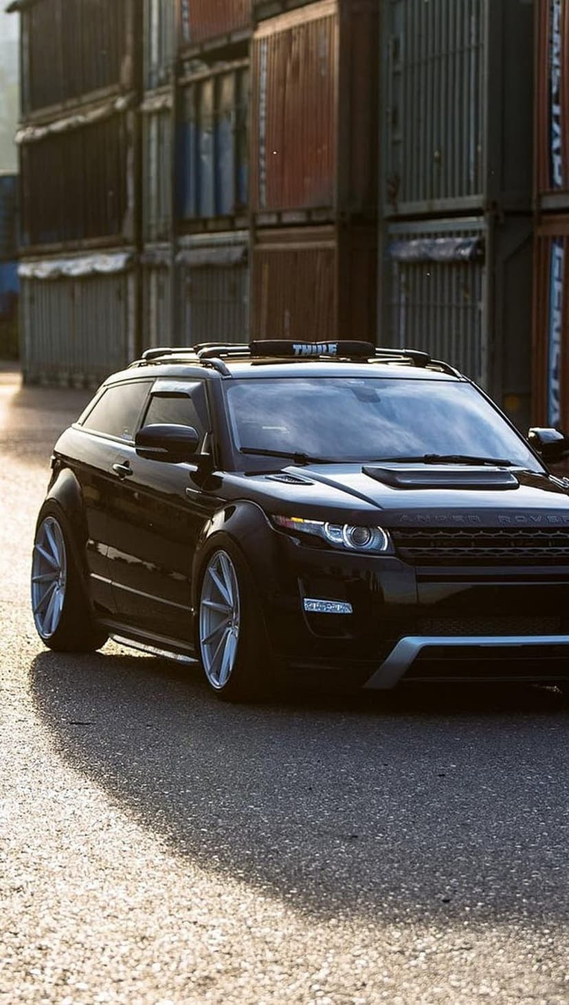 Black Range Rover Evoque SUV with silver wheels and a roof rack parked near stacked industrial shipping containers.