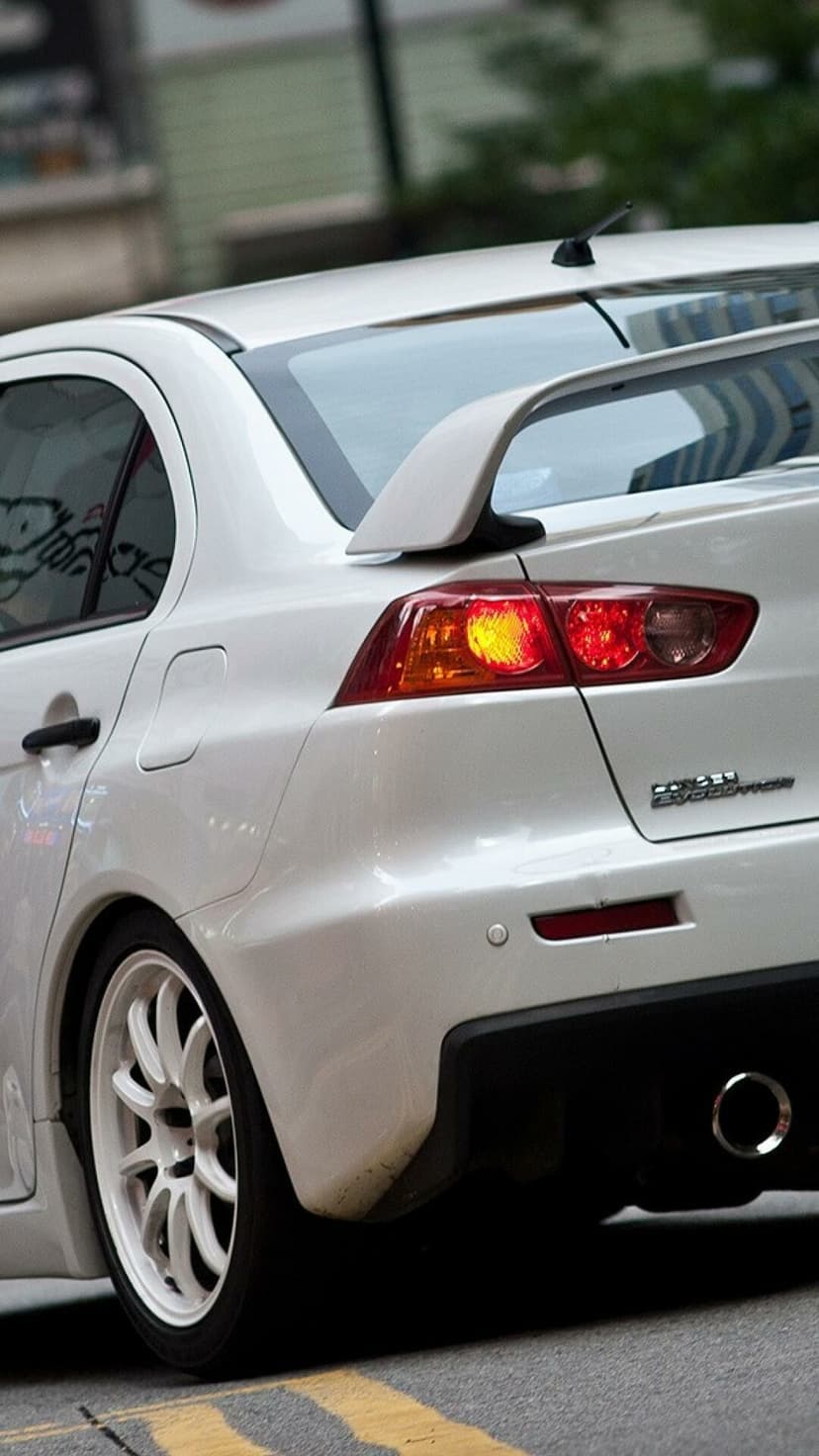 Rear view of a white Mitsubishi Lancer Evolution X with white rims, a large spoiler, and glowing taillights.