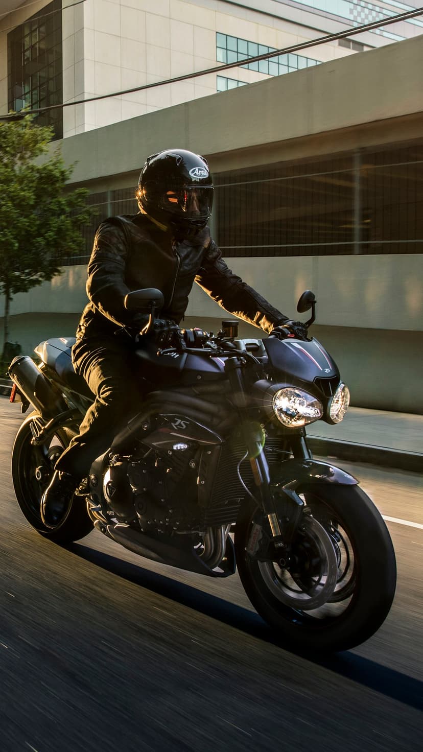 Motorcyclist in a leather jacket and helmet riding through an urban street at sunset with long shadows.