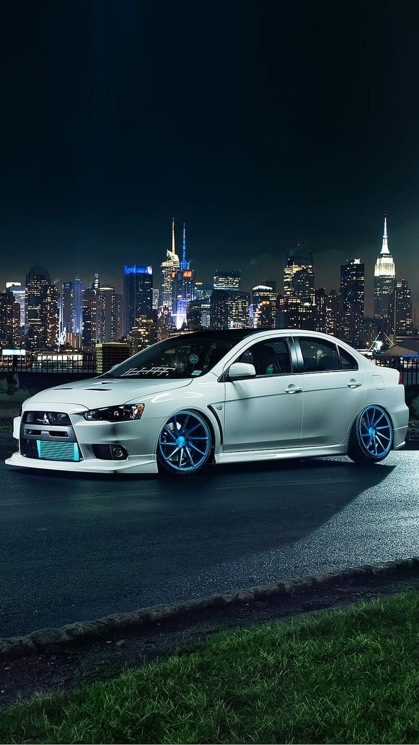White Lancer Evolution X with blue neon lights parked on damp asphalt before a bokeh city skyline at night.