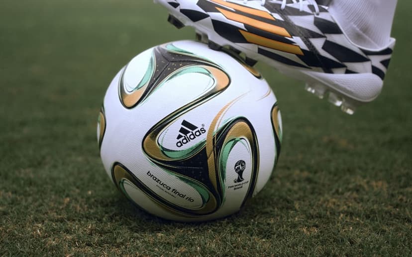 Adidas Brazuca soccer ball and a gold-patterned white cleat sit on green grass blades in a close-up view.