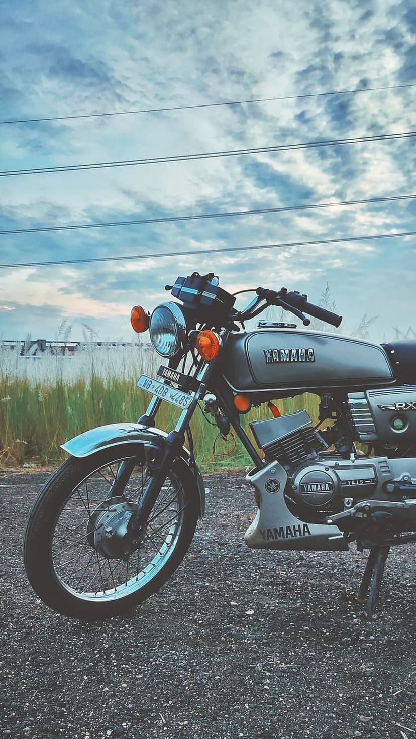 Silver Yamaha RX 100 motorcycle parked on a gravel path against a cloudy sunset sky in a rural field setting.