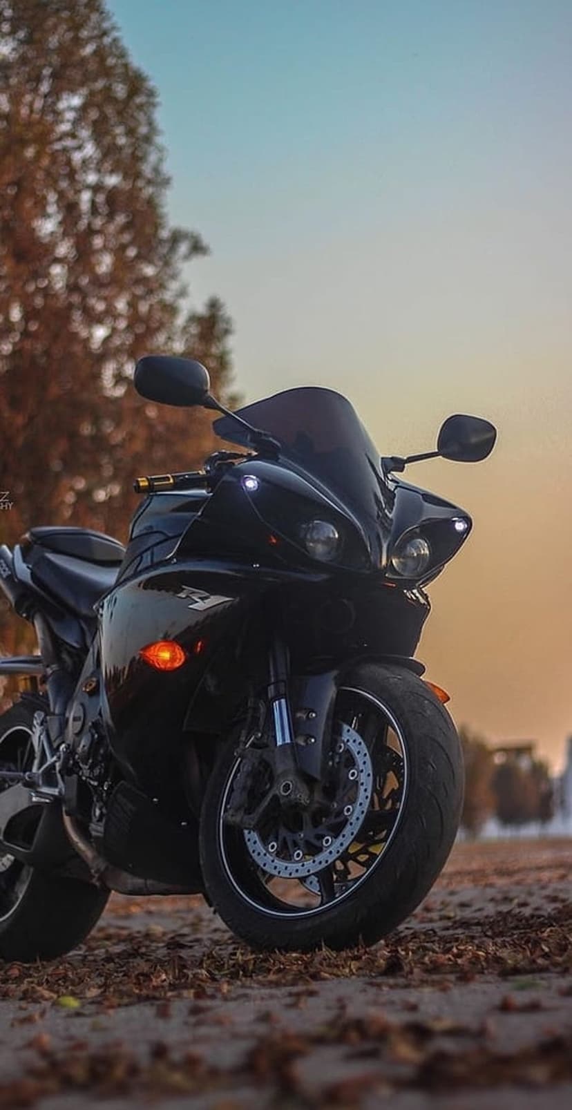 Yamaha R1 black sportbike parked on autumn leaves with glowing headlights against a dusk sunset gradient sky.