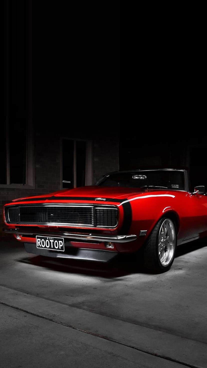 Red 1969 Chevrolet Camaro convertible with black racing stripes and chrome accents parked on a dark urban street.