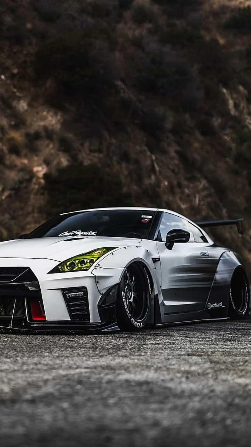 White Nissan GT-R with widebody kit, large rear wing, and custom wheels parked on a textured asphalt road surface.