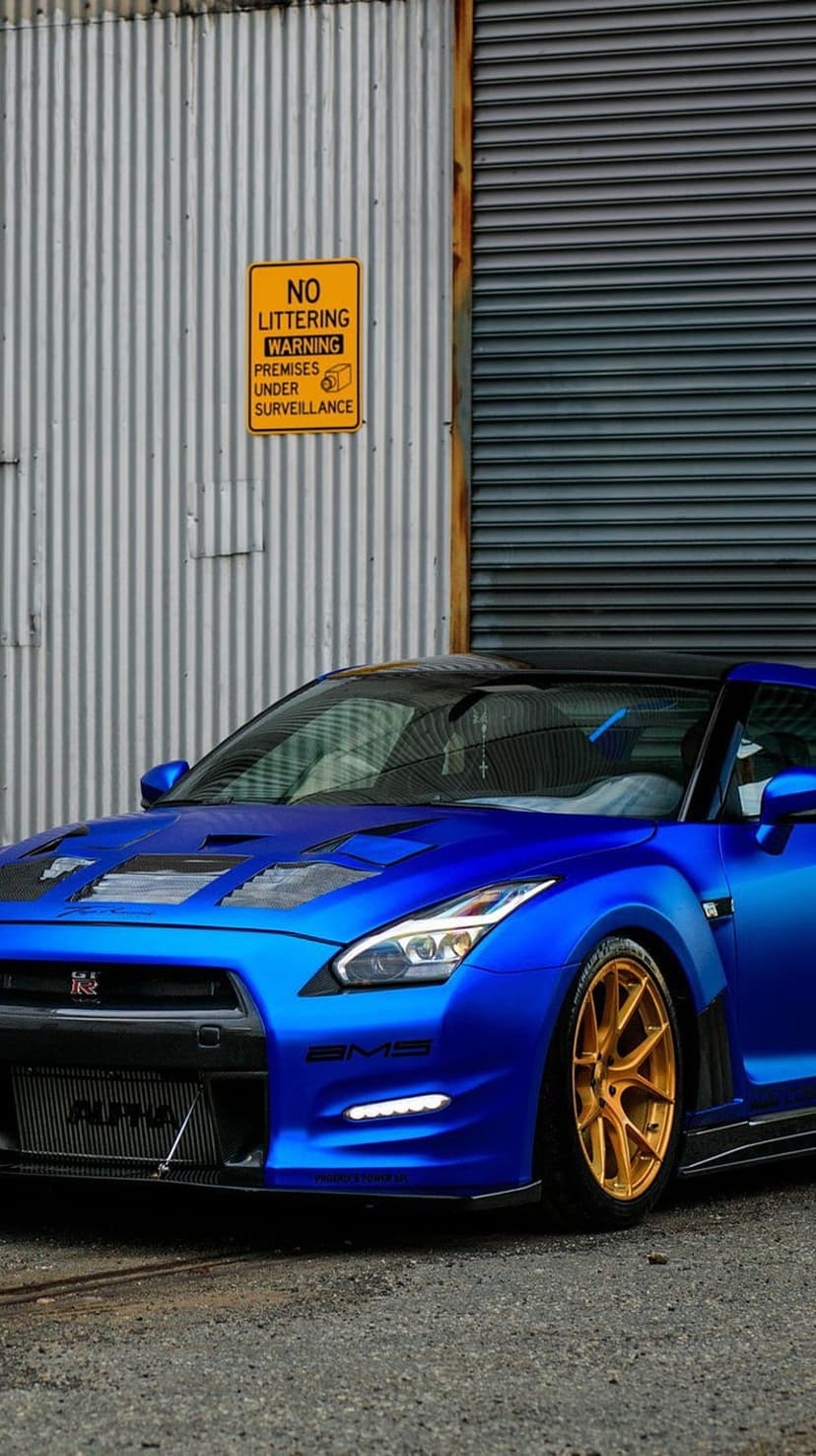 Nissan GT-R with matte blue paint and gold rims parked before a corrugated metal wall with a no littering sign.