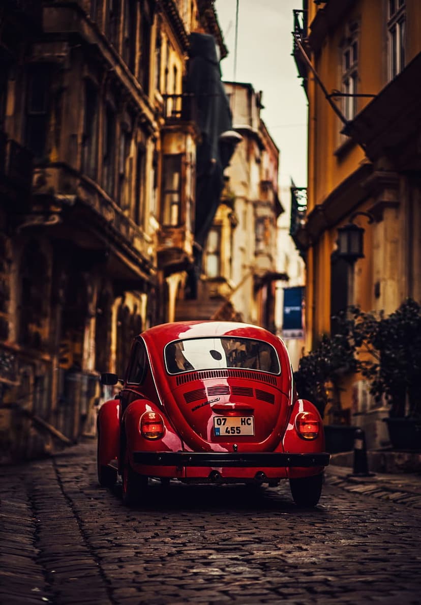 Red Volkswagen Beetle parked on a narrow cobblestone street between old European buildings in soft lighting.