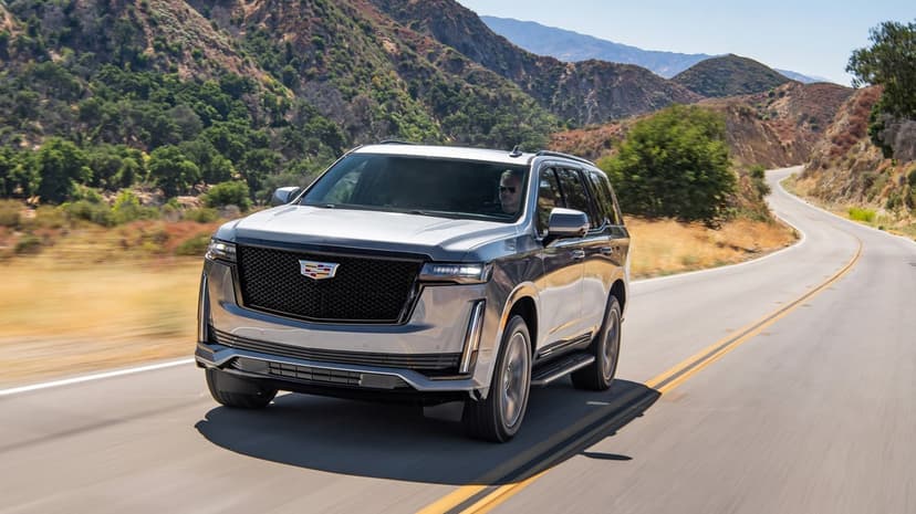 Silver Cadillac Escalade driving on a winding mountain road under a clear blue sky during daylight.