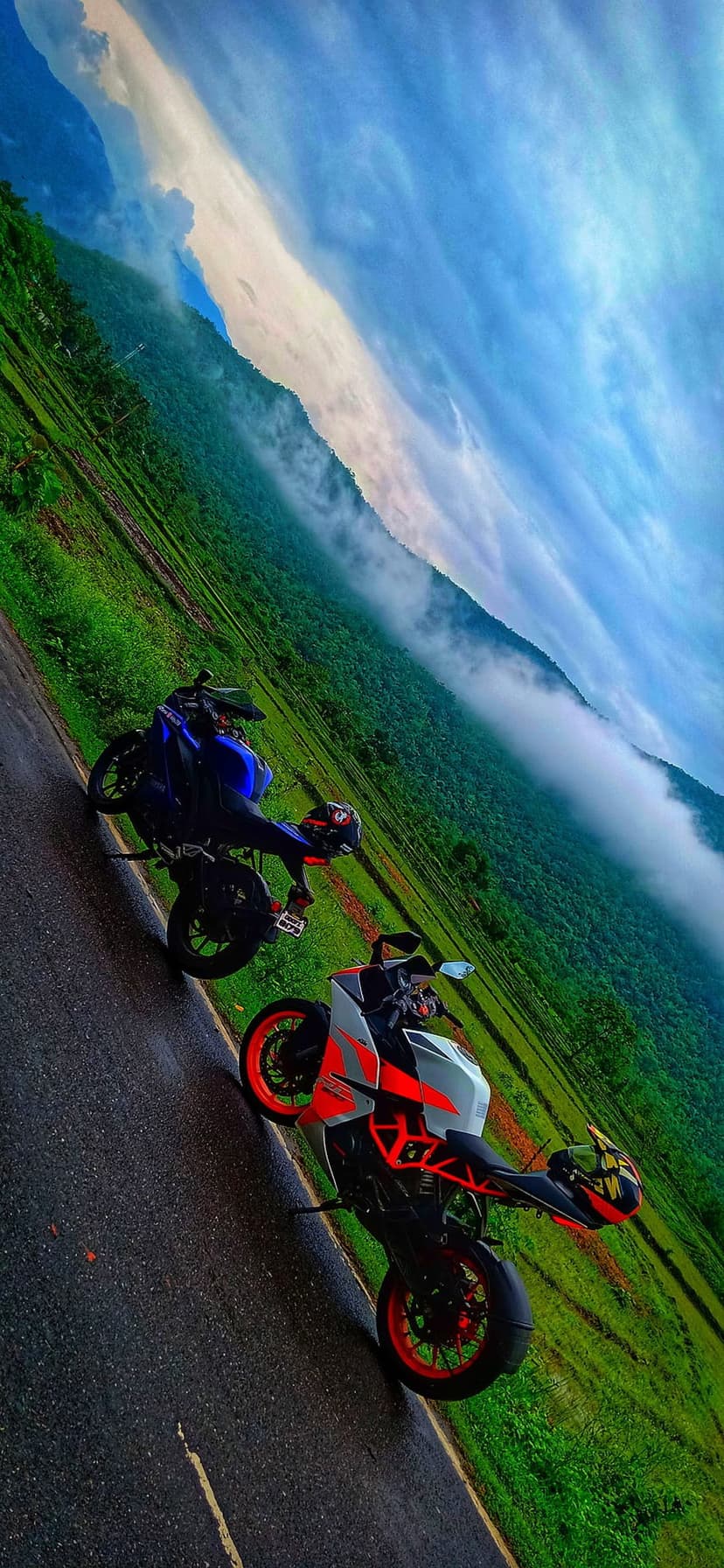 Blue and orange sport motorcycles parked on a wet mountain road overlooking misty green terraced hills under clouds.