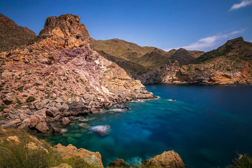 Layered rocky cliffs descend into clear blue ocean water under a bright sunlit sky with submerged rocks visible.