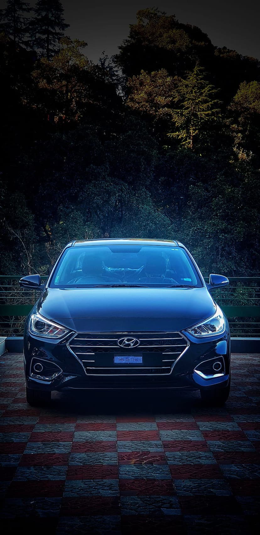 Dark blue Hyundai Verna sedan parked on checkered tiles with dense green trees under a moody, overcast sky.