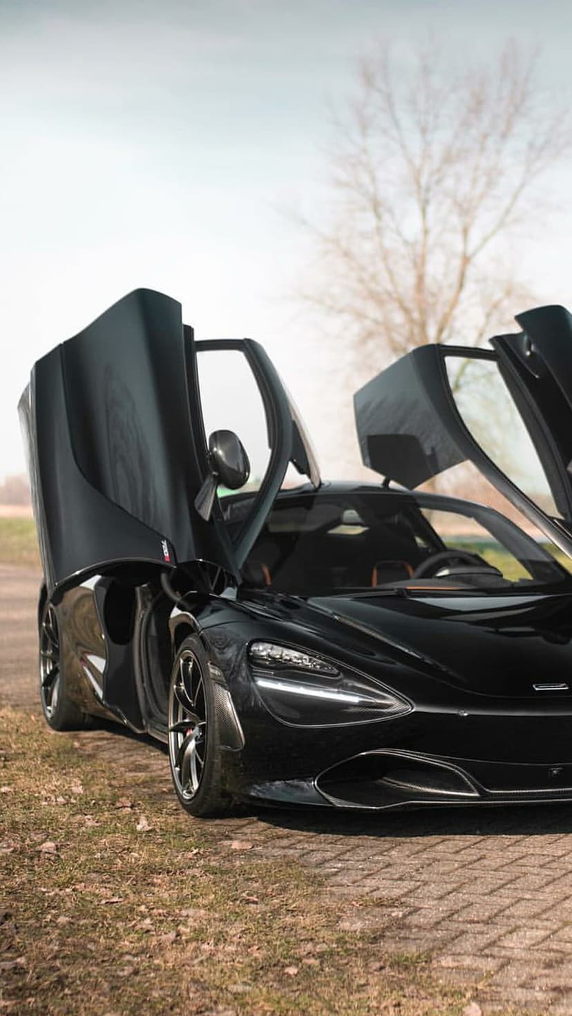 Black McLaren 720S supercar with butterfly doors open upward in a field with dry grass and a leafless tree.