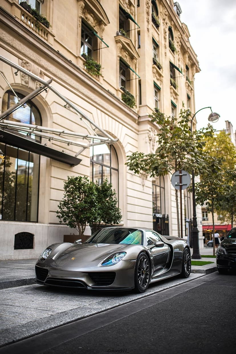 Silver Porsche 918 Spyder parked on a cobblestone street in front of a grand Parisian stone building with trees.