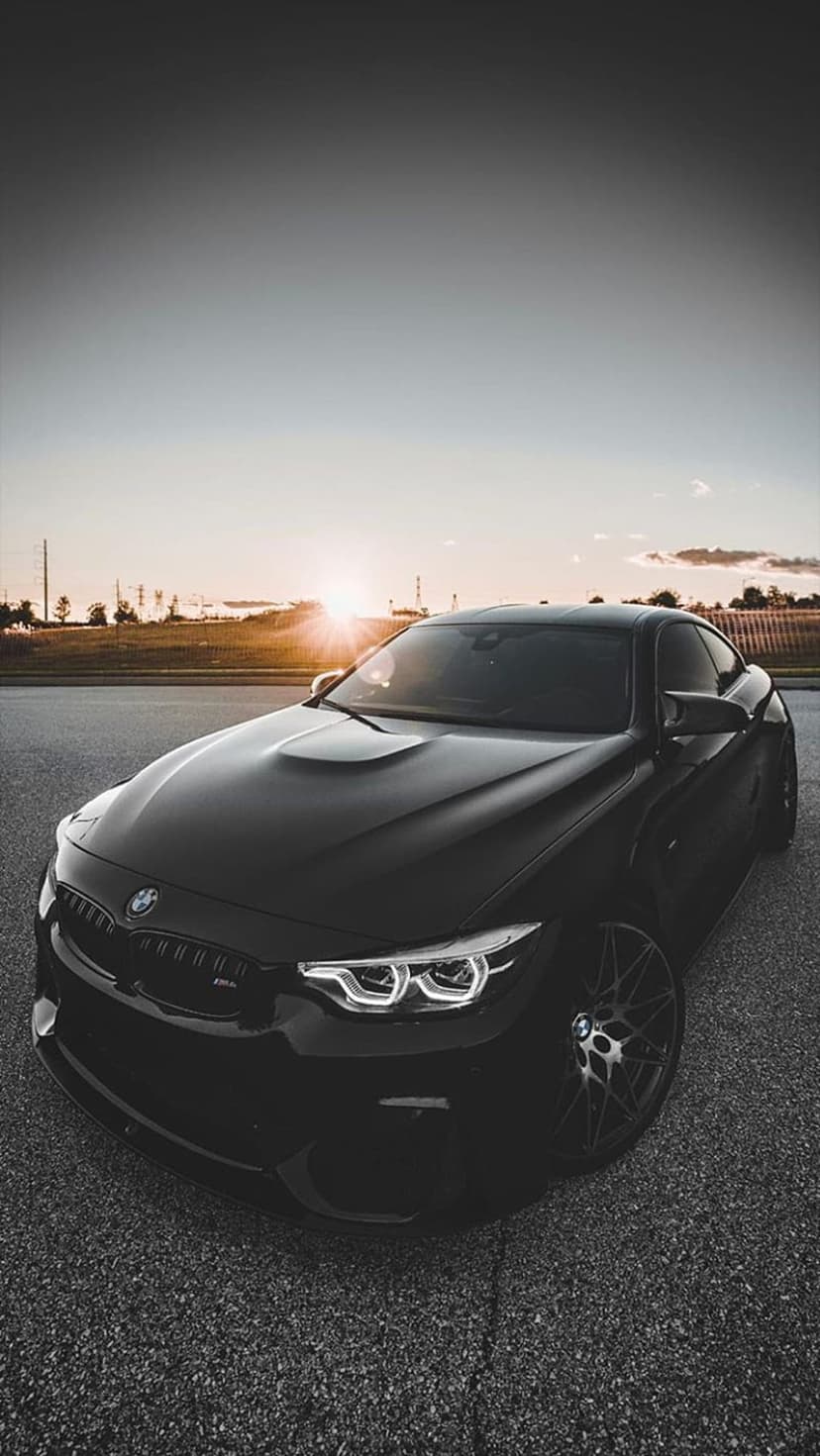 Black BMW M4 sports car parked on asphalt with warm sunset light reflecting off its hood and LED headlights.