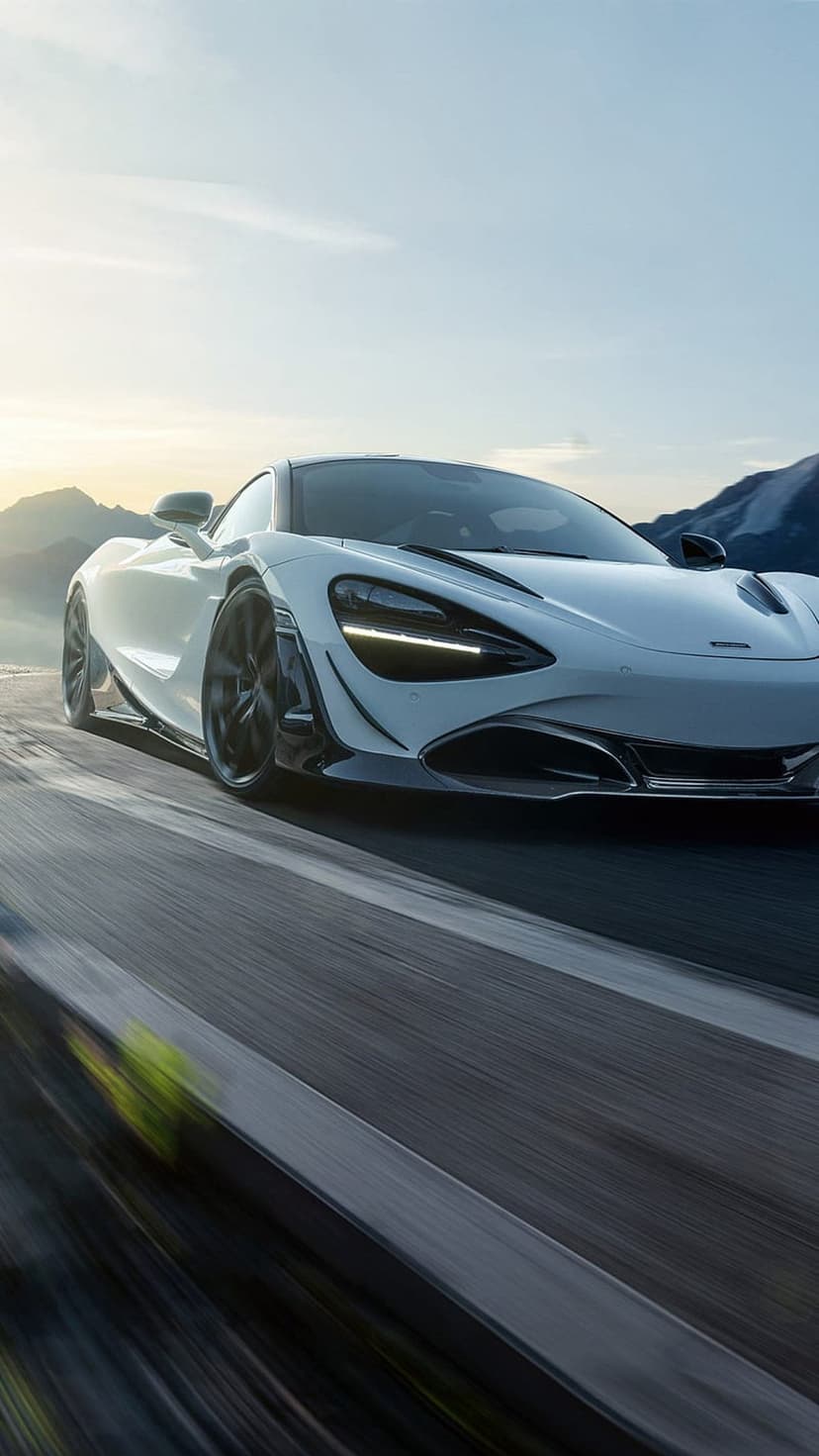 White McLaren 720S supercar speeds through a winding mountain road with motion blur under a bright, clear sky.