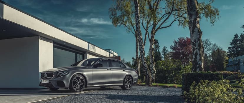 Matte silver Mercedes-Benz E-Class parked on gravel by a modern white house under a clear blue sky.