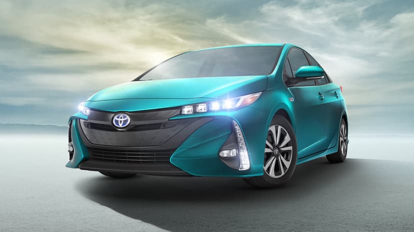 Cyan Toyota Prius Prime hybrid car with glowing LED headlights on a misty flat ground under a cloudy sky.