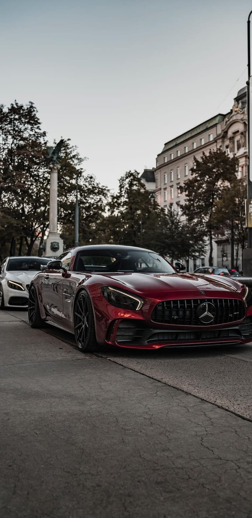 Red Mercedes-AMG GT parked on cracked asphalt in front of historic buildings and trees on a city street.
