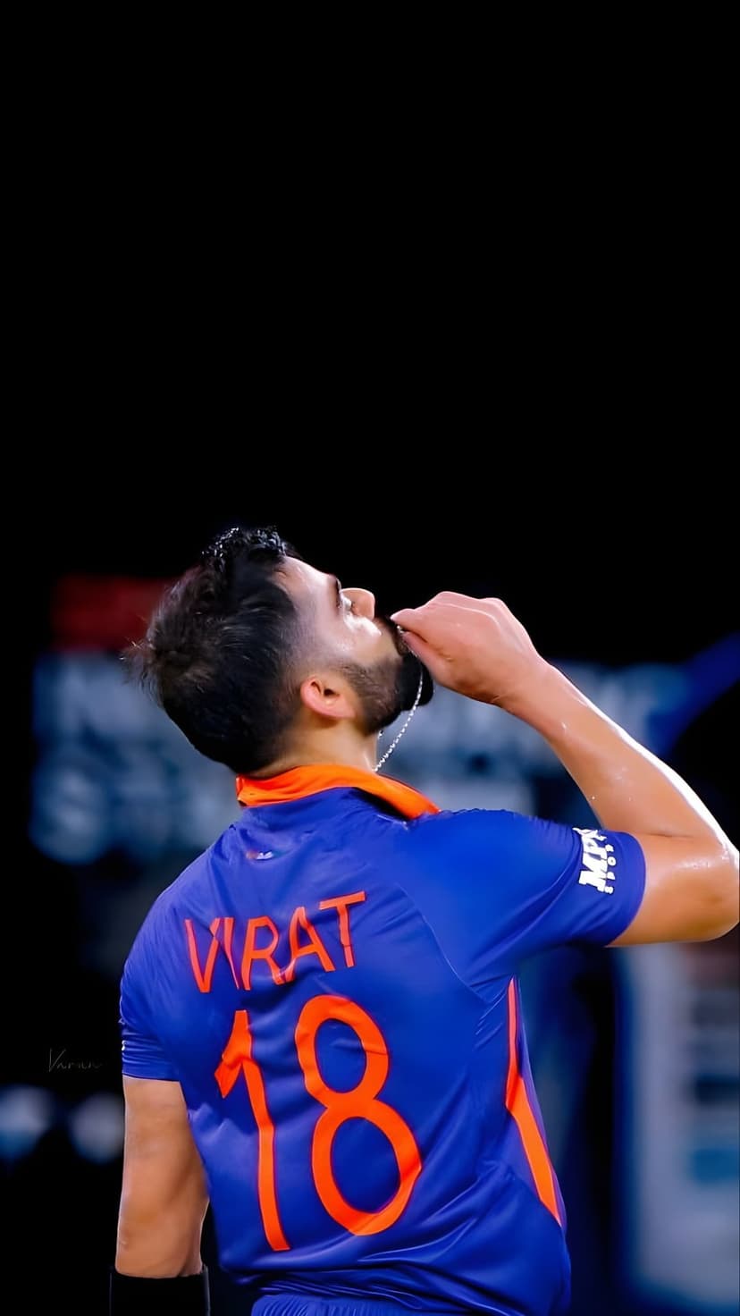 Virat Kohli kisses his bat wearing a blue India jersey with orange number 18 against a dark, moody background.