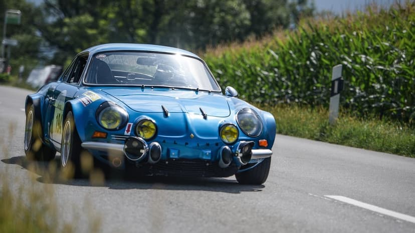 Blue vintage Alpine A110 rally car with yellow headlights driving on a paved road next to a green cornfield.