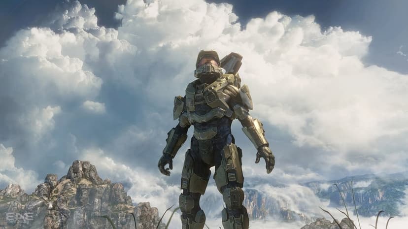 Master Chief in green armor stands on a rocky mountain peak against a backdrop of massive white clouds and sky.
