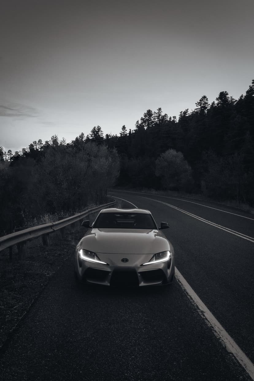 Toyota Supra with glowing headlights on a winding forest road in a dramatic black and white landscape.