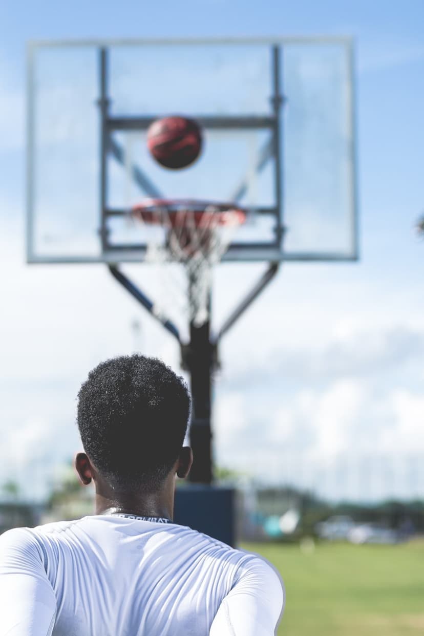 African American man in a white shirt watches a basketball fly toward an outdoor hoop under a clear blue sky.