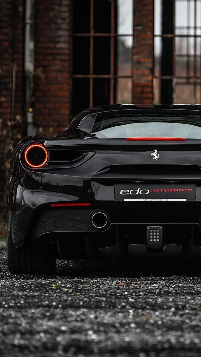 Ferrari 488 Competition rear view with circular taillights and silver emblem parked on gravel near a brick wall.