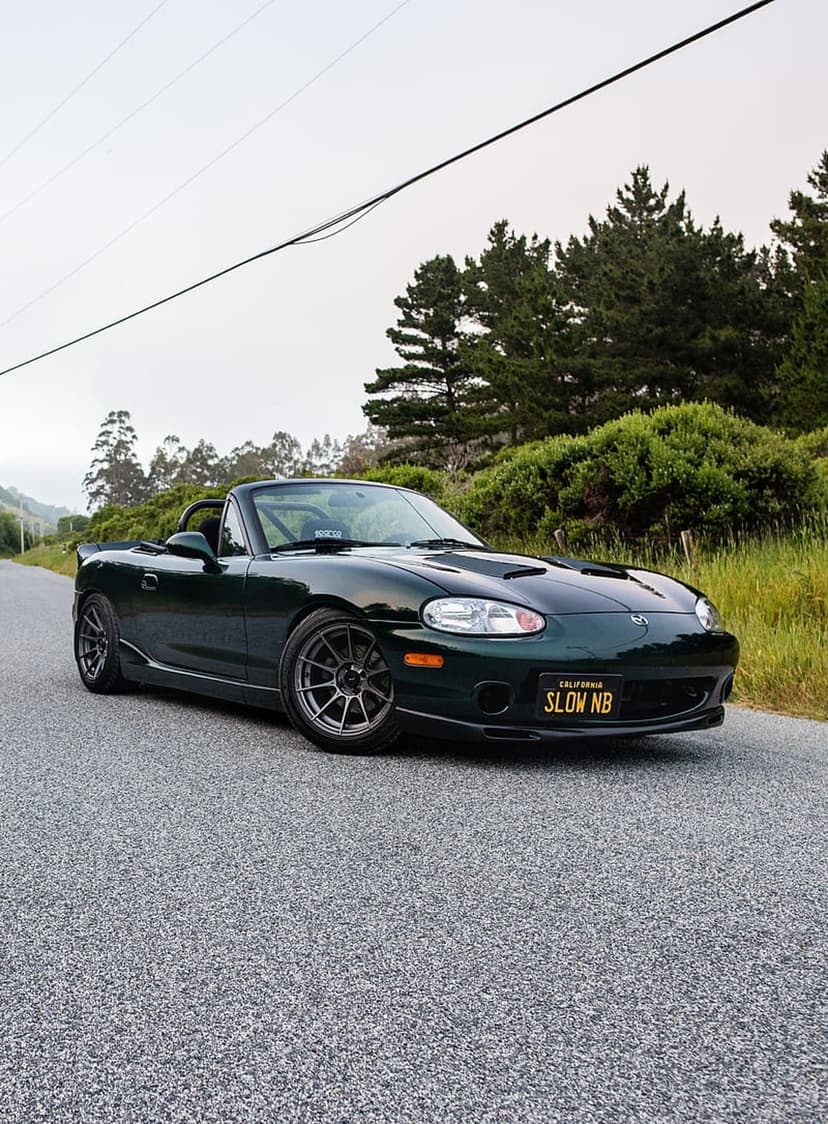 Dark green Mazda Miata NB convertible with custom wheels parked on an asphalt road among lush green trees.