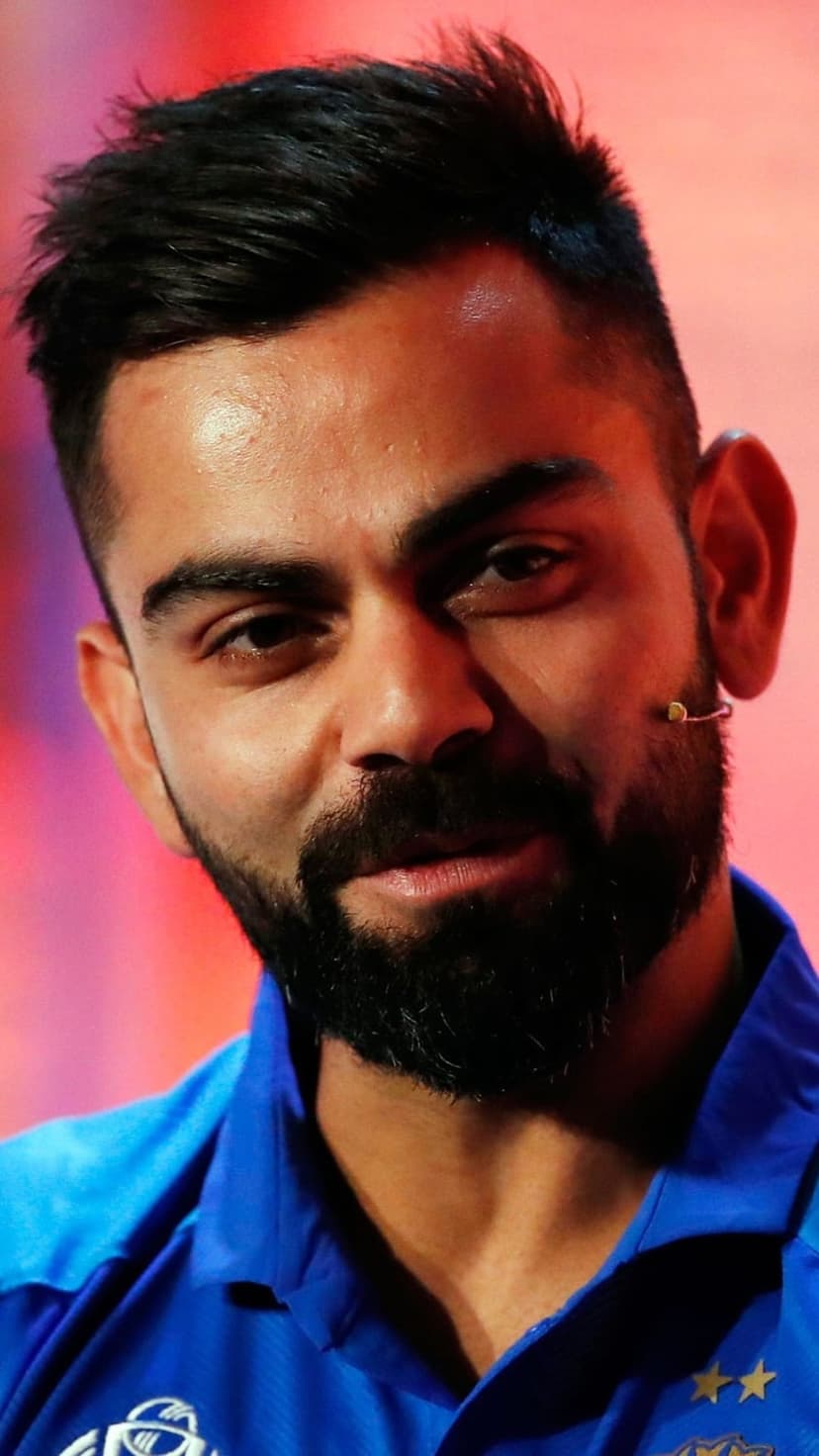 Virat Kohli with a trimmed beard and styled hair wears a blue shirt against a blurred red and pink background.