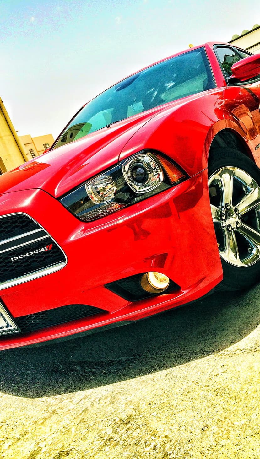 Red Dodge Charger front grill and aggressive headlights shown from a low angle on sunny asphalt under a blue sky.