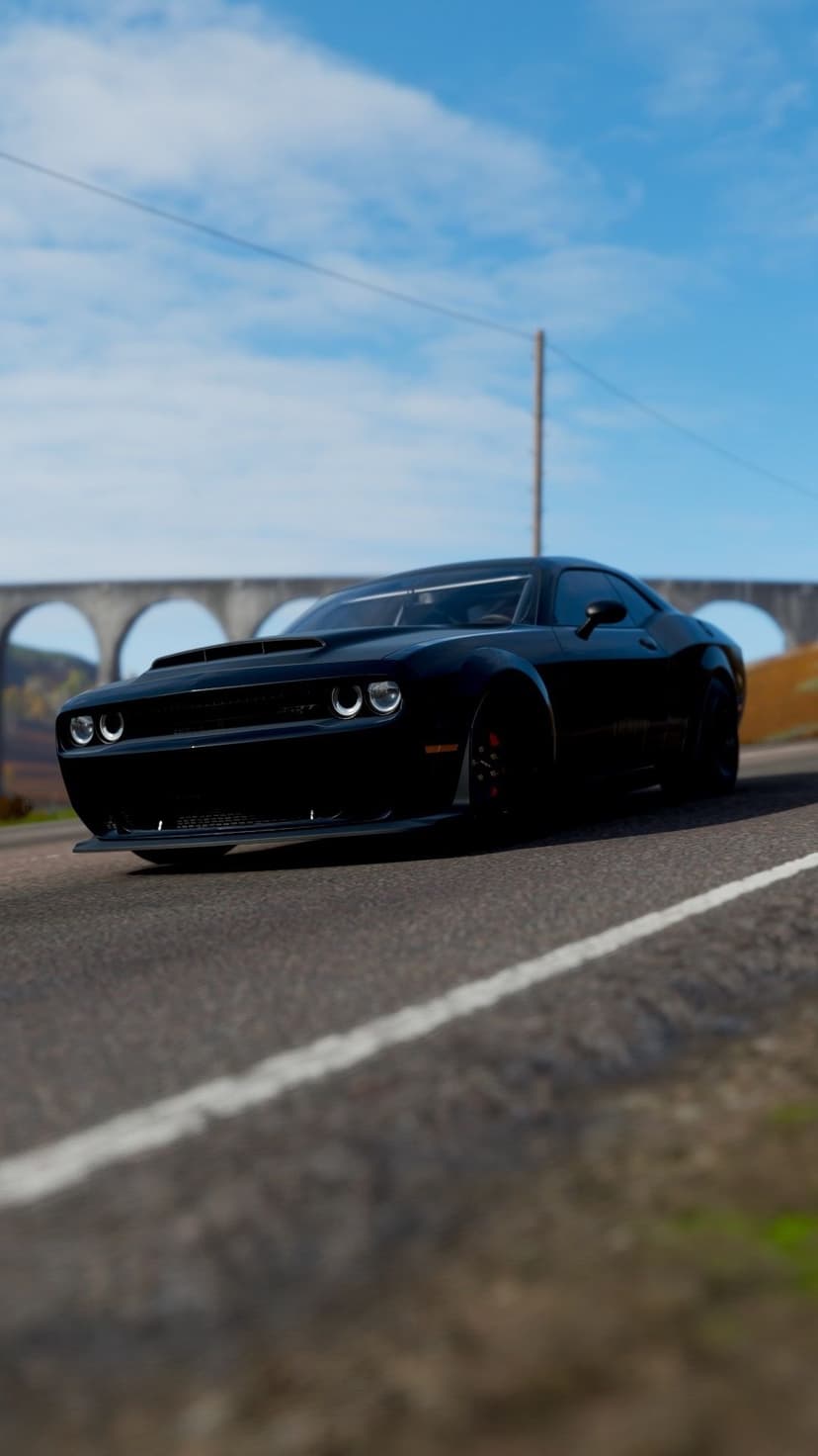 Black muscle car on a paved road with a stone arch bridge and blue sky in the background under bright daylight.