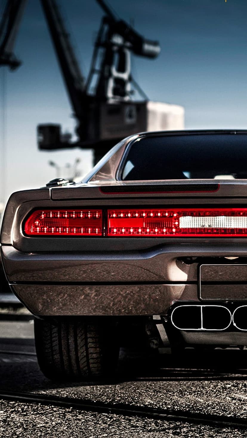 Muscle car rear with red LED taillights and dual exhausts parked near a large industrial crane at a shipping dock.