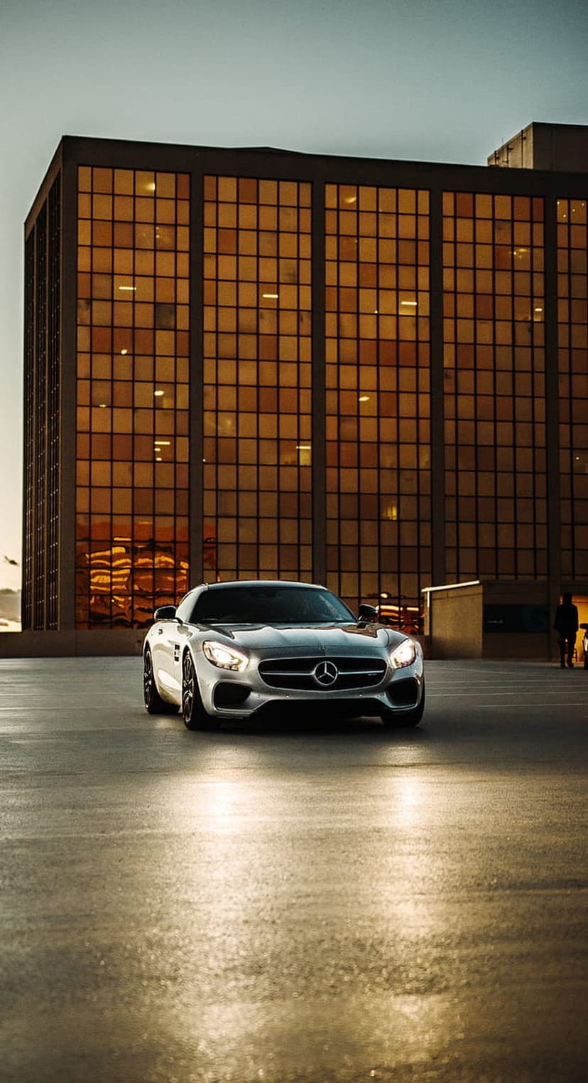 Silver Mercedes AMG GT on wet pavement reflecting the golden glow of a modern skyscraper in an urban setting.