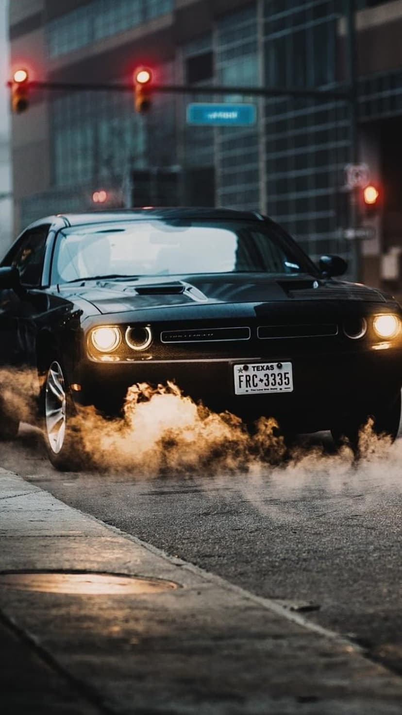 Black Dodge Challenger spins its tires, creating a thick smoke cloud on an urban street at night with city lights.