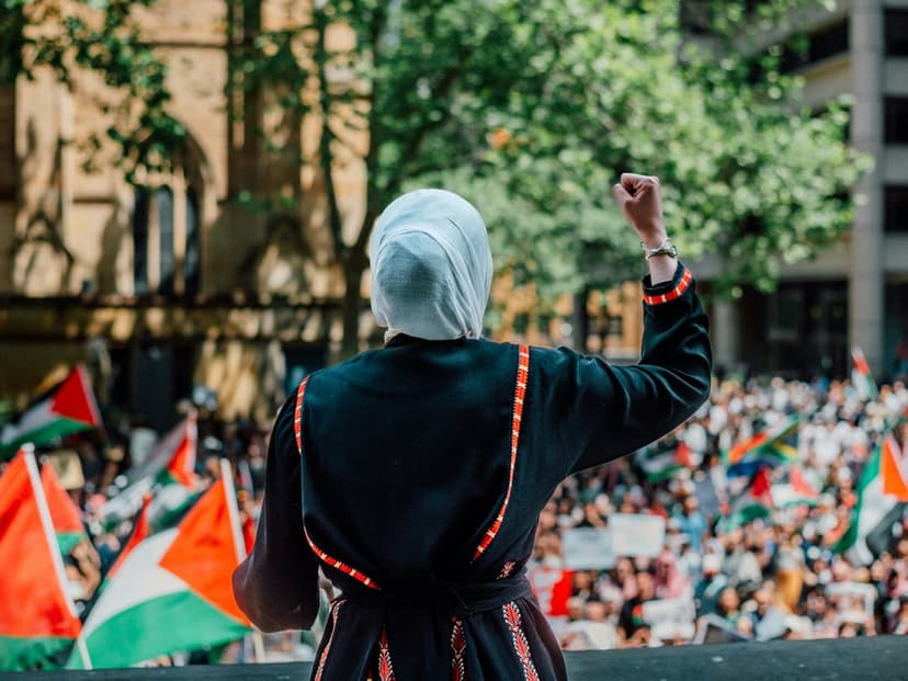 Woman in a white hijab raises her fist amid a crowd waving Palestinian flags at an outdoor protest rally.