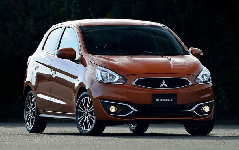 Orange Mitsubishi Mirage hatchback parked on asphalt with glowing headlights and a dark forest background.