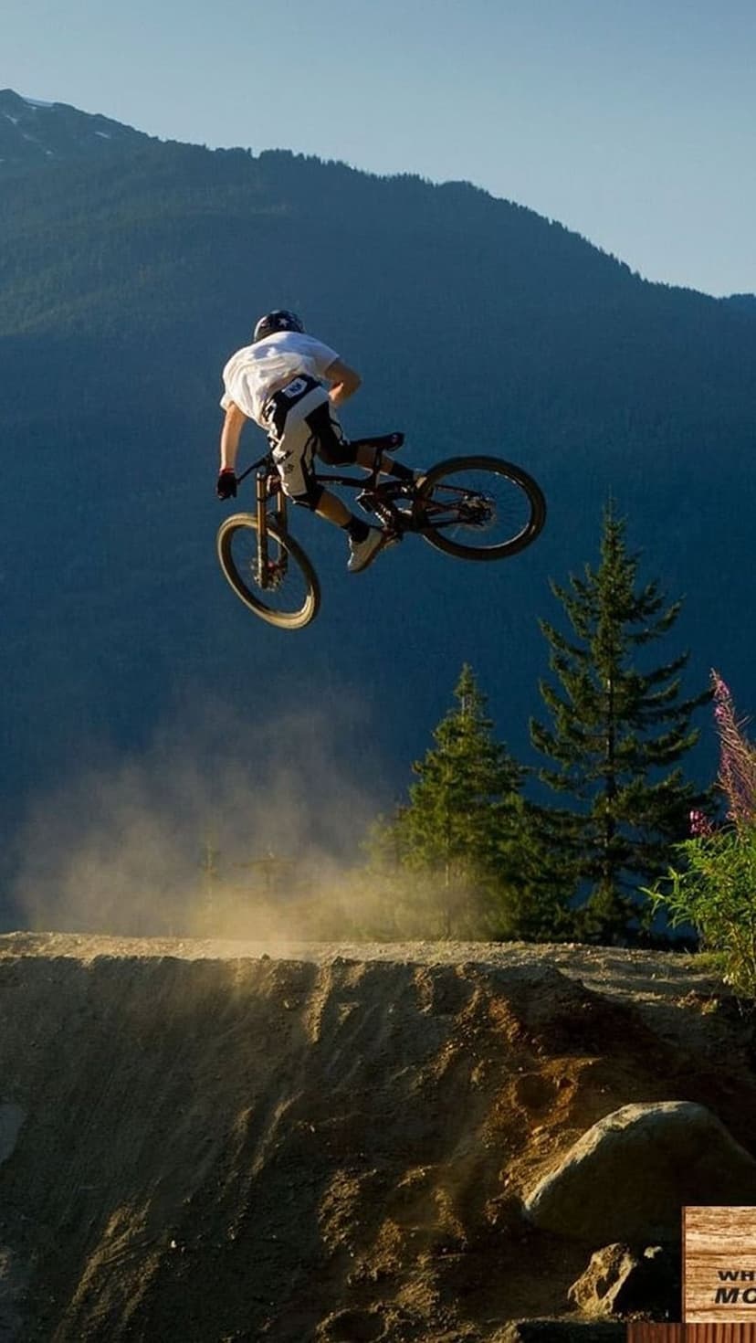 Mountain biker in protective gear jumping mid-air over a dirt track with pine trees and peaks in the background.