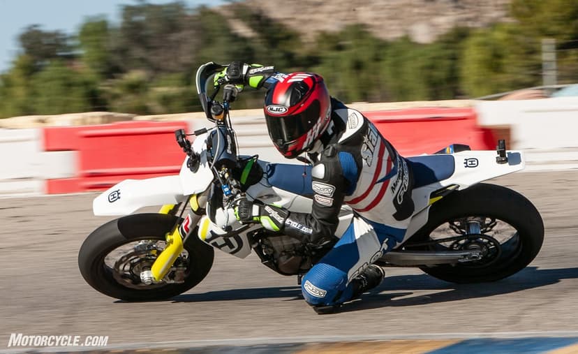 Motorcyclist in a racing suit leaning deep into a turn on a white Husqvarna supermoto bike at a racetrack.