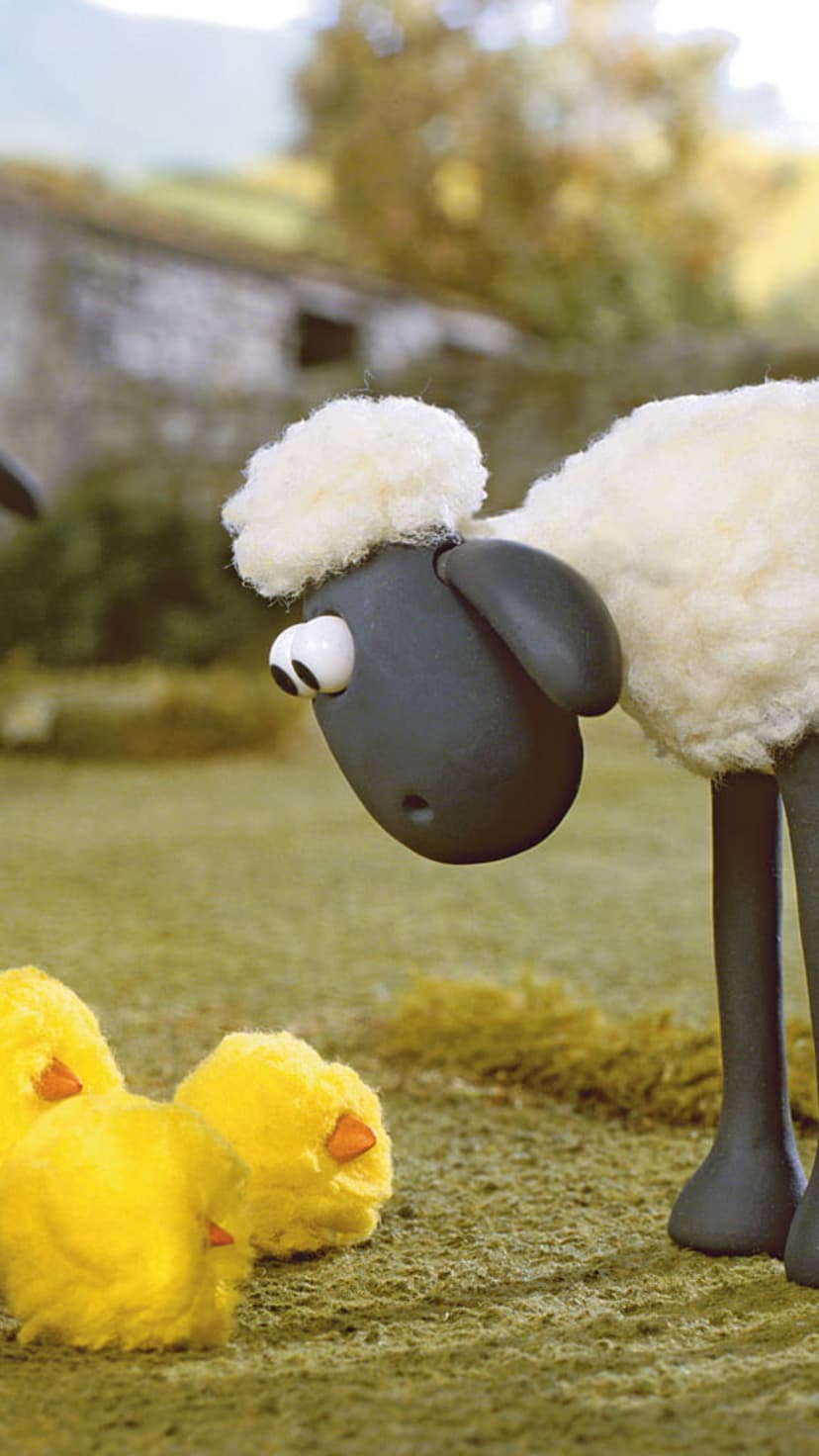 Shaun the Sheep stares down at three tiny yellow chicks on a grassy farmyard with a stone barn in the distance.