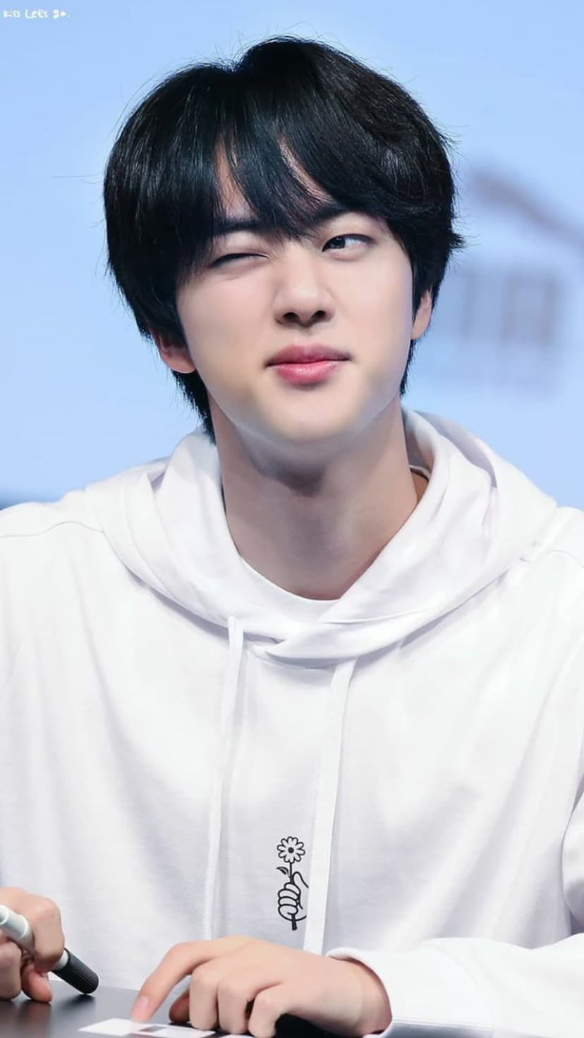 Jin from BTS winks playfully while wearing a white hoodie with a flower graphic against a soft blue background.