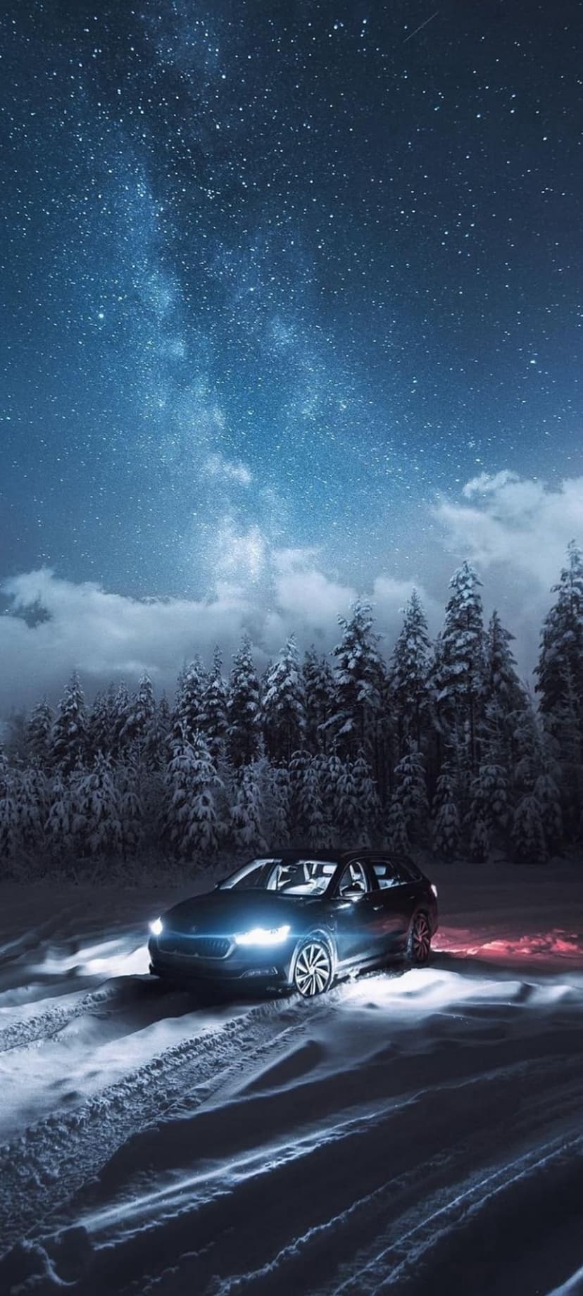 Dark car driving on a snowy road through pine trees under a starry night sky with the visible Milky Way galaxy.